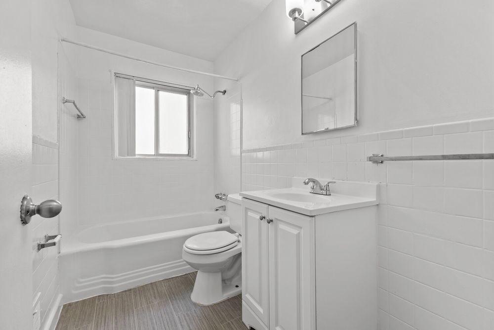 Clean white bathroom with subway tile, soaking tub, toilet, and vanity with mirror on wood-look floors.
