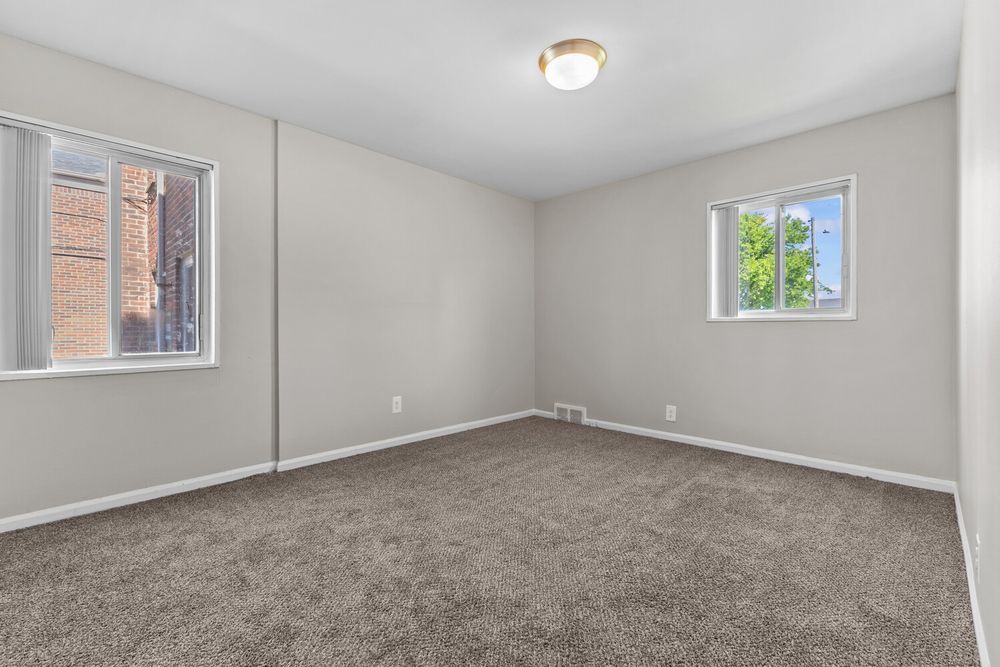 Empty carpeted bedroom with two windows, neutral gray walls, and a ceiling light fixture.