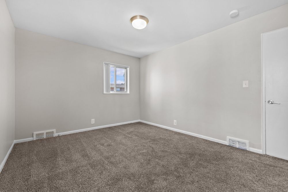 Empty bedroom with brown carpet, light gray walls, small window, and ceiling light fixture.