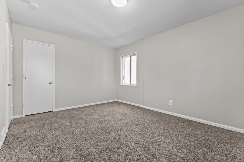 Empty bedroom with gray carpet, white walls, a single window, and a white door in a clean, modern home.