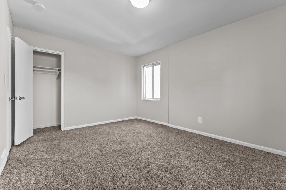 Empty bedroom with gray carpet, white walls, open closet door, and a single window providing natural light.