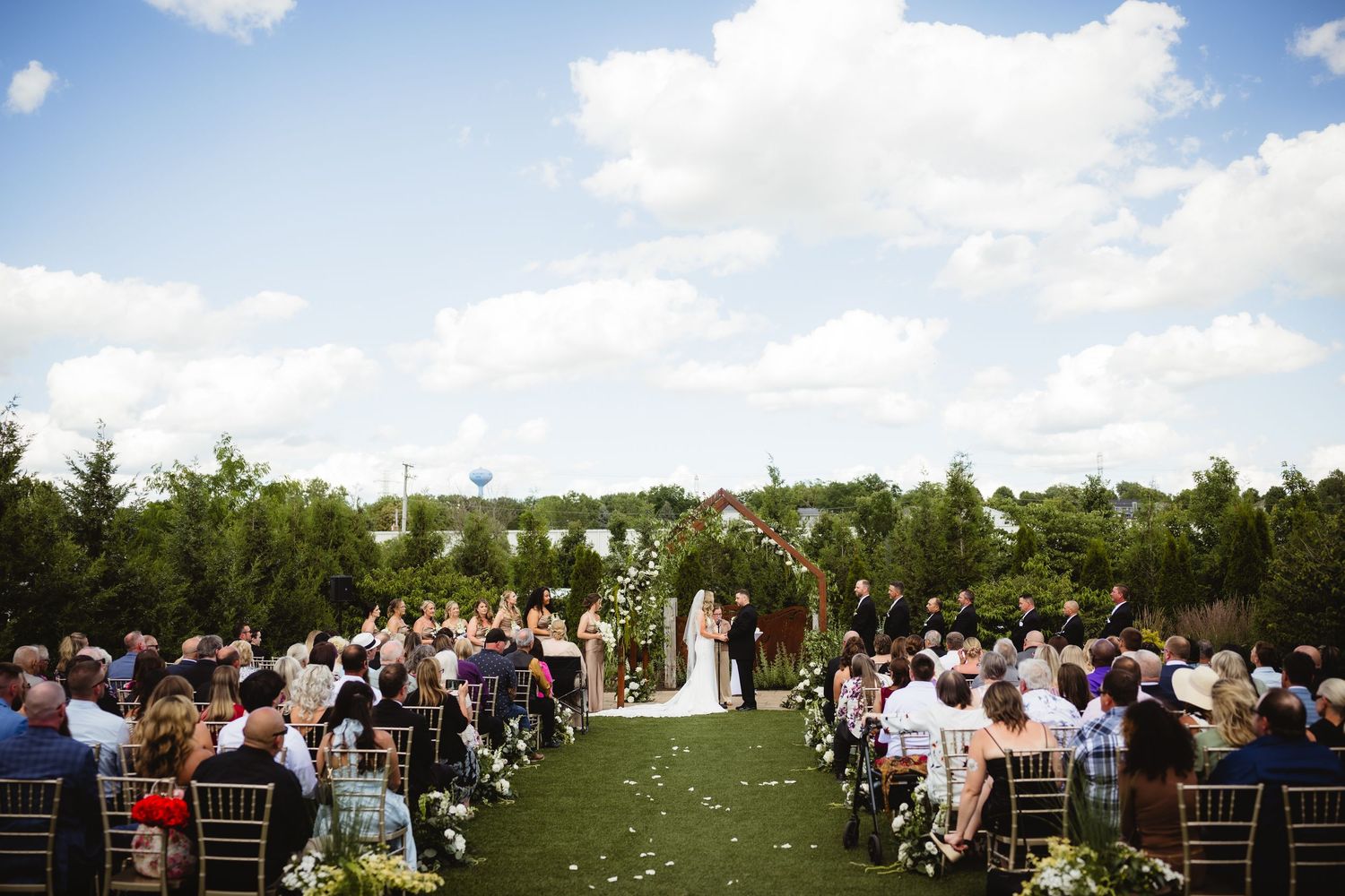 Outdoor wedding ceremony on green lawn with floral arch, seated guests, and blue sky with clouds.