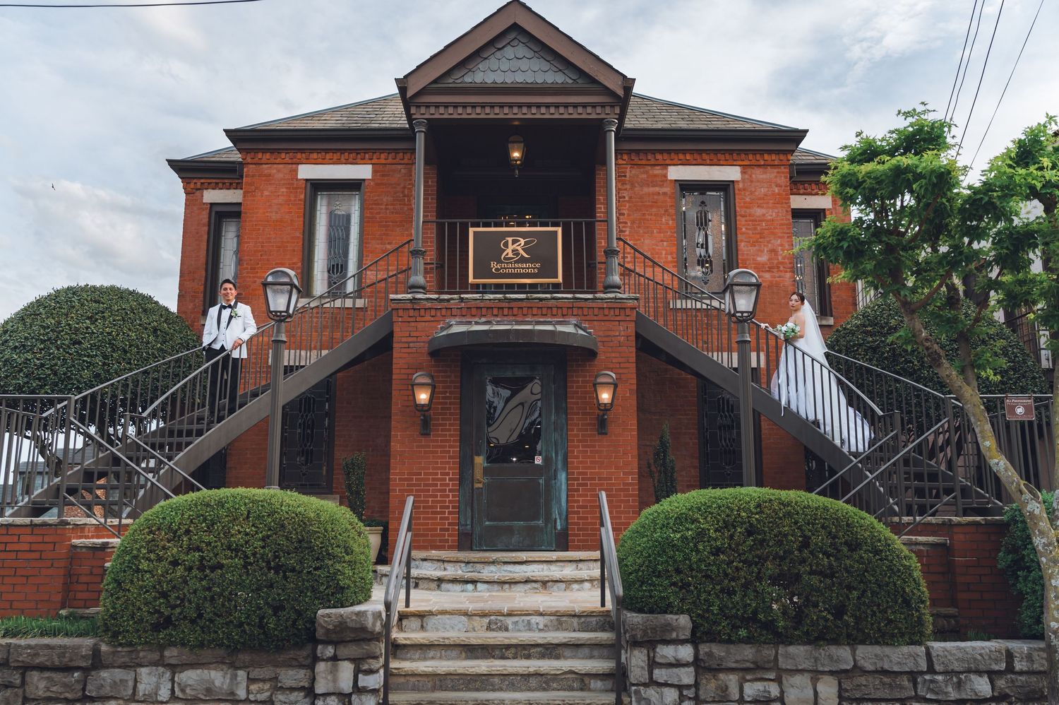 Elegant red brick Victorian venue with dual staircases, manicured hedges, and ornate ironwork railing under cloudy skies.
