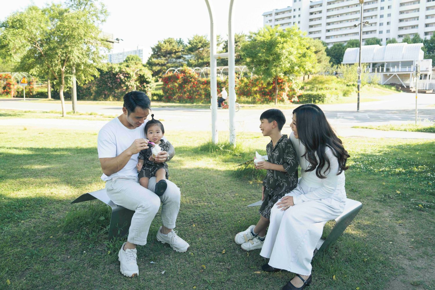 Family of four enjoying a sunny day at the park, sitting on benches with children eating snacks outdoors.