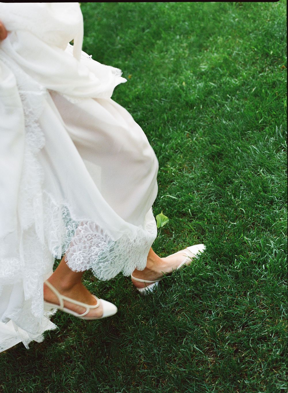 Bride in white lace wedding dress and ivory heels walking on green grass.