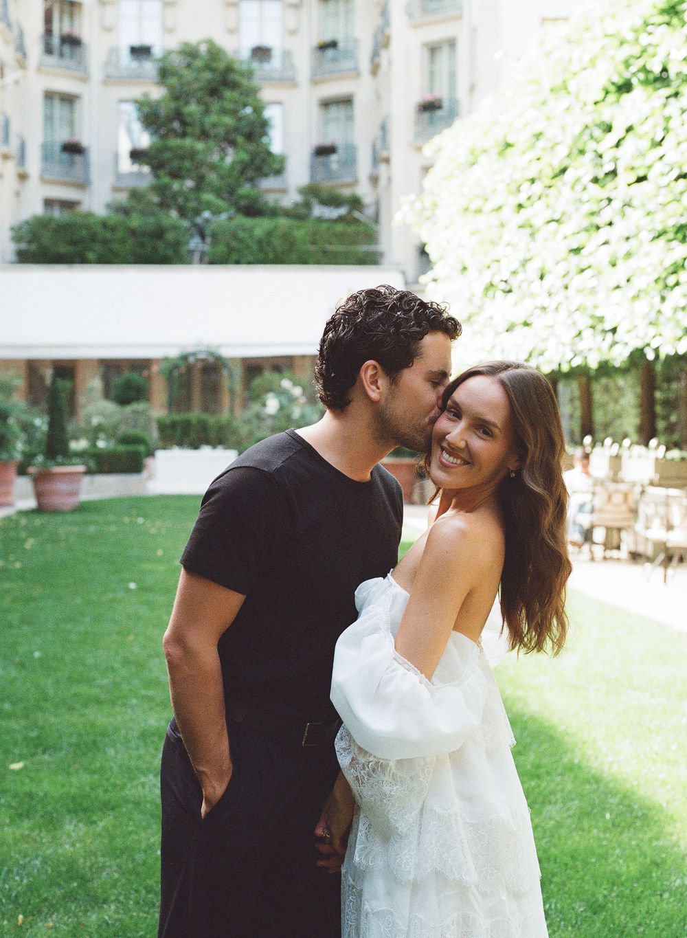 Couple embracing in lush garden courtyard, man kissing woman's cheek as she smiles, elegant hotel backdrop.