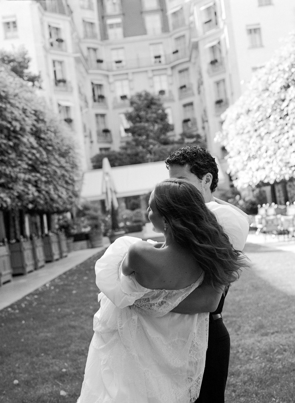 Black and white photo of a couple embracing in a Parisian courtyard, bride in off-shoulder gown, elegant building backdrop.