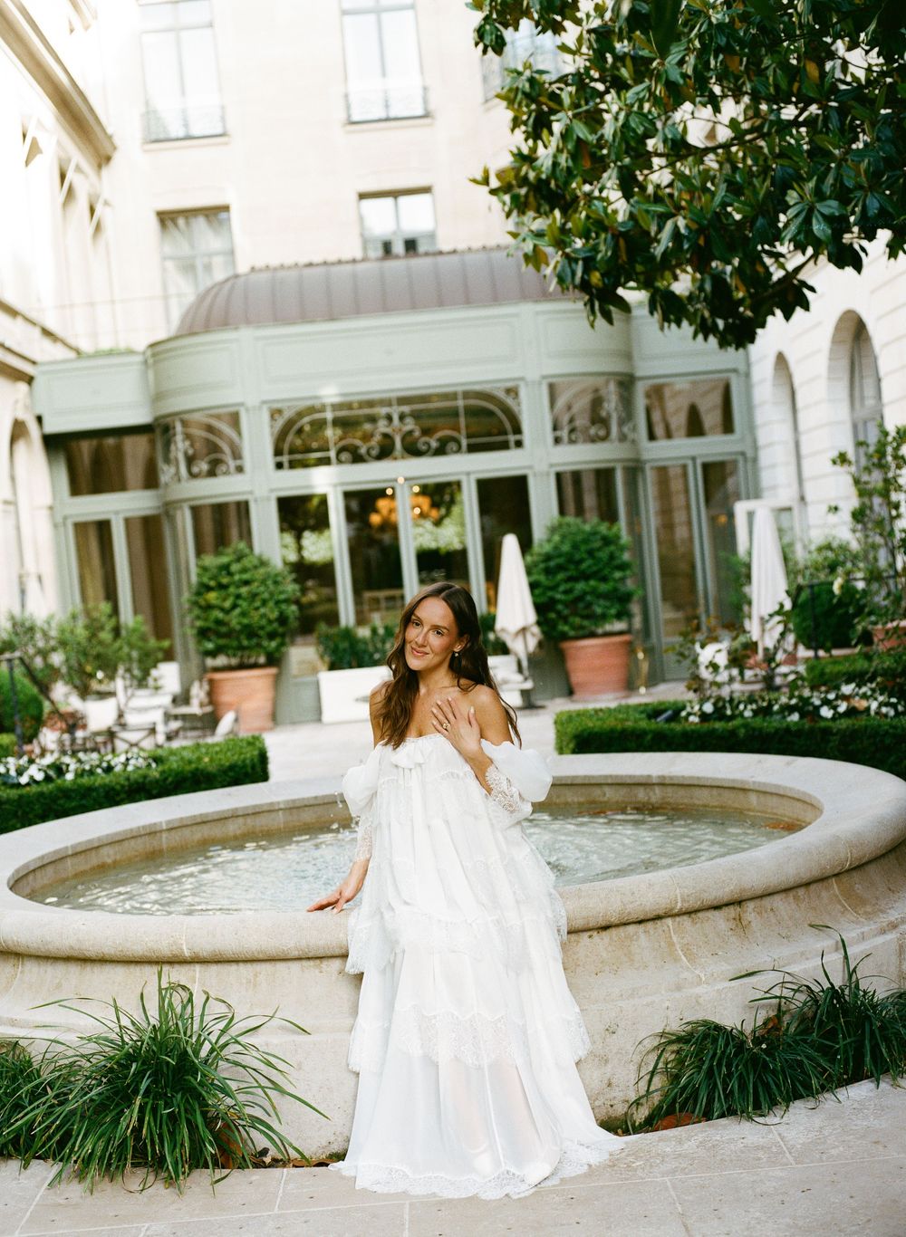 Bride in white off-shoulder tiered gown poses by stone fountain in elegant hotel courtyard garden.