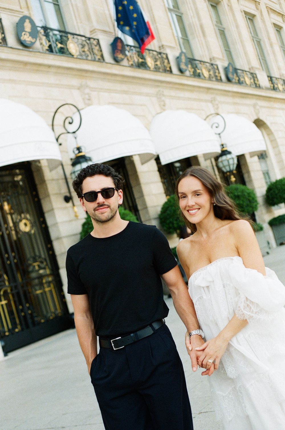 Couple holding hands smiling in front of elegant Parisian building, man in black outfit, woman in white off-shoulder dress.