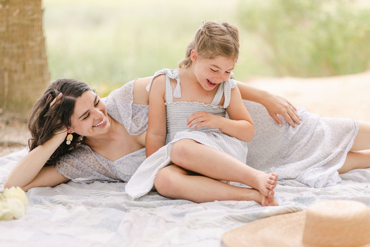 A mother and young daughter laugh together on a blanket outdoors, both wearing matching gray dresses.