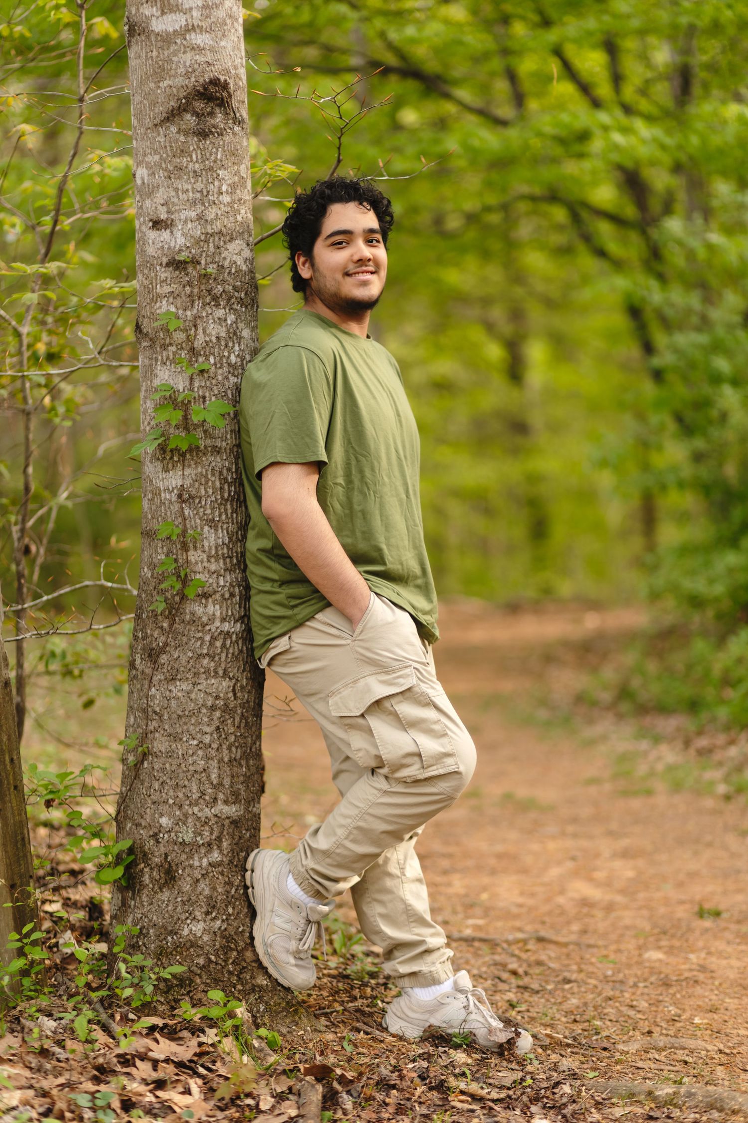 Dark-haired senior boy wearing a green shirt rests against a tree against a fresh spring backdrop, by Charlottesville family photographer.
