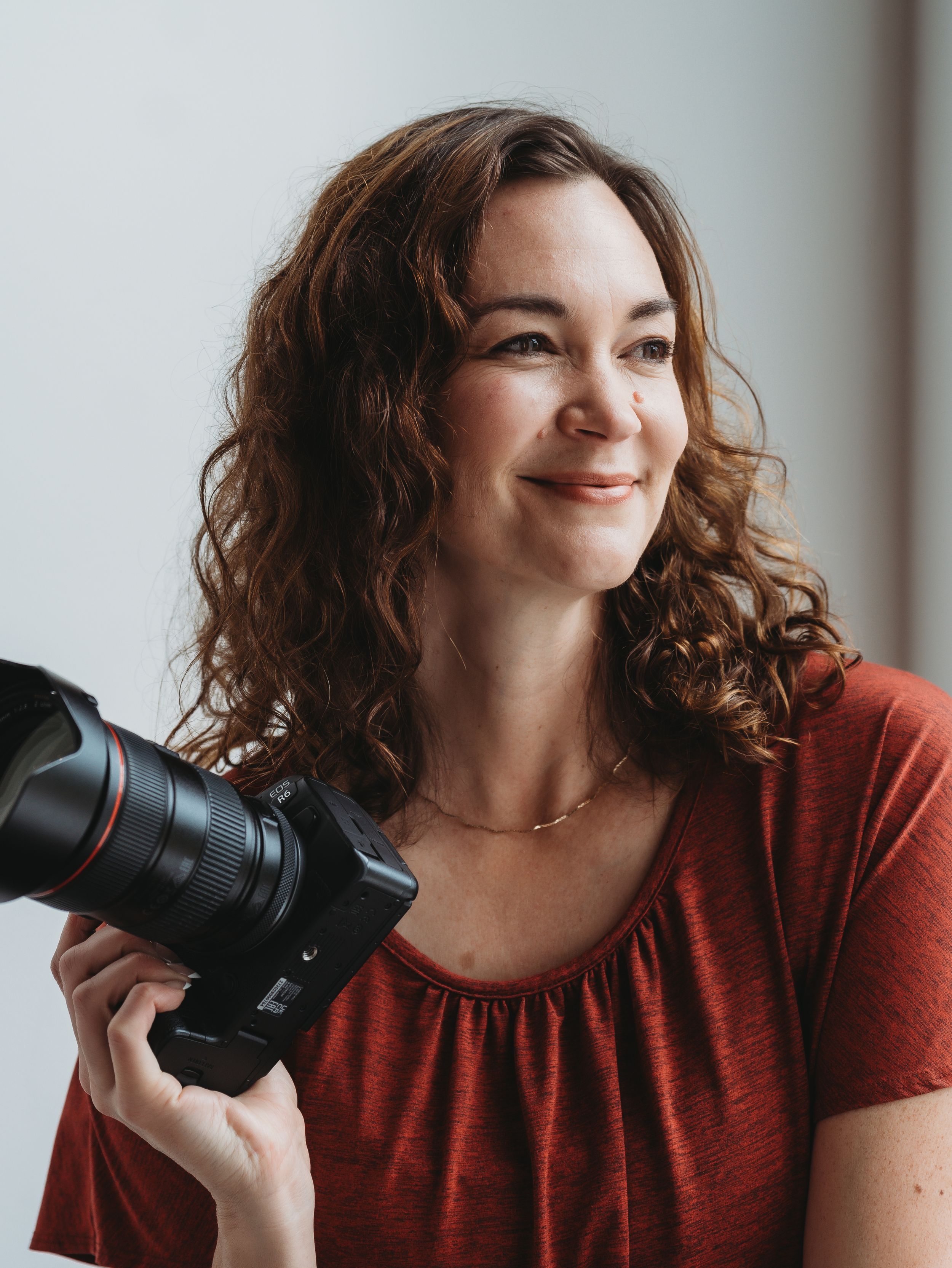 Black and white portrait of a woman leaning against a wall holding a camera, smiling softly in elegant dark attire.