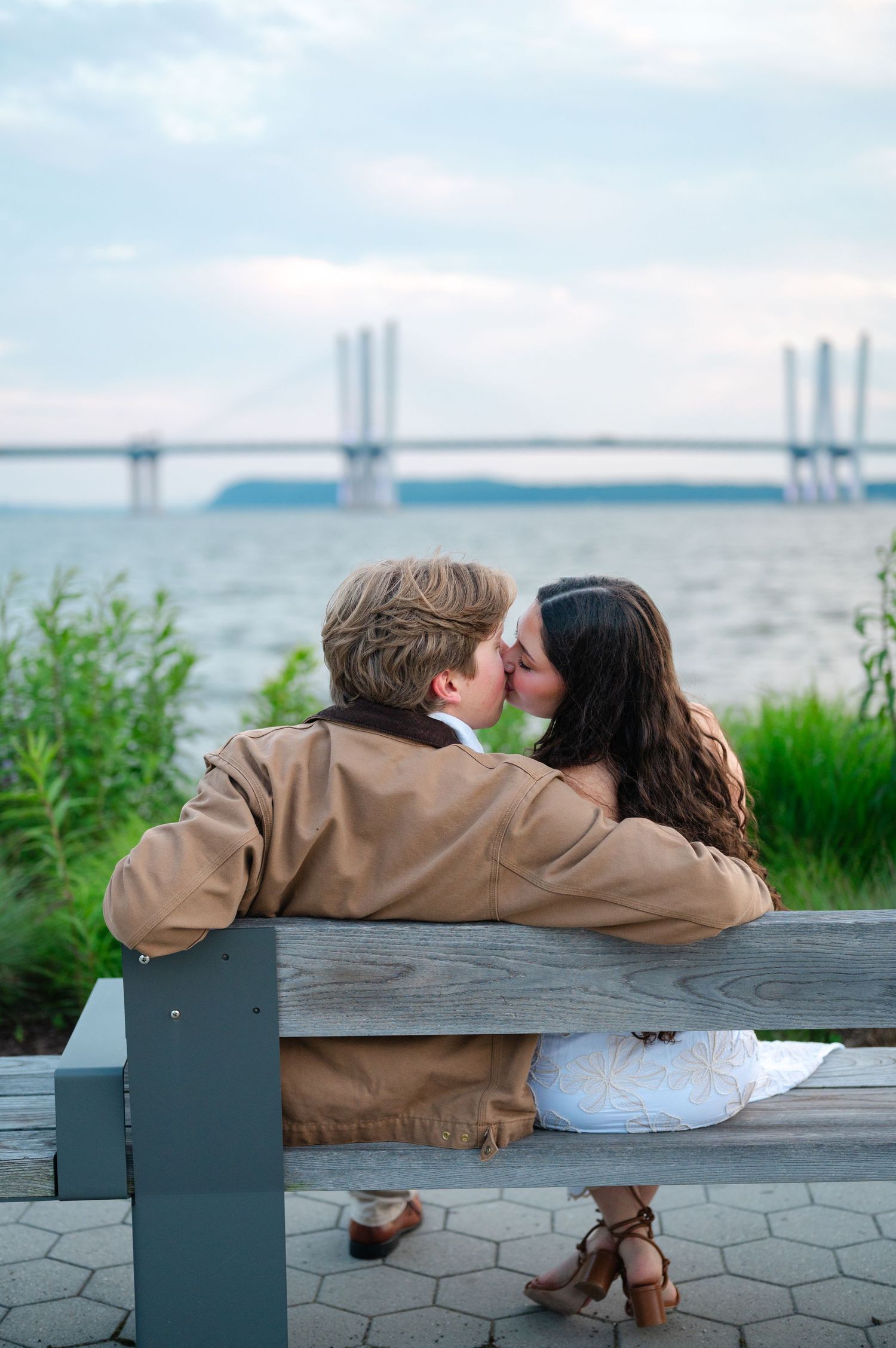 Couple sharing a romantic kiss on a wooden bench overlooking the Hudson River with a bridge in the background after their proposal.