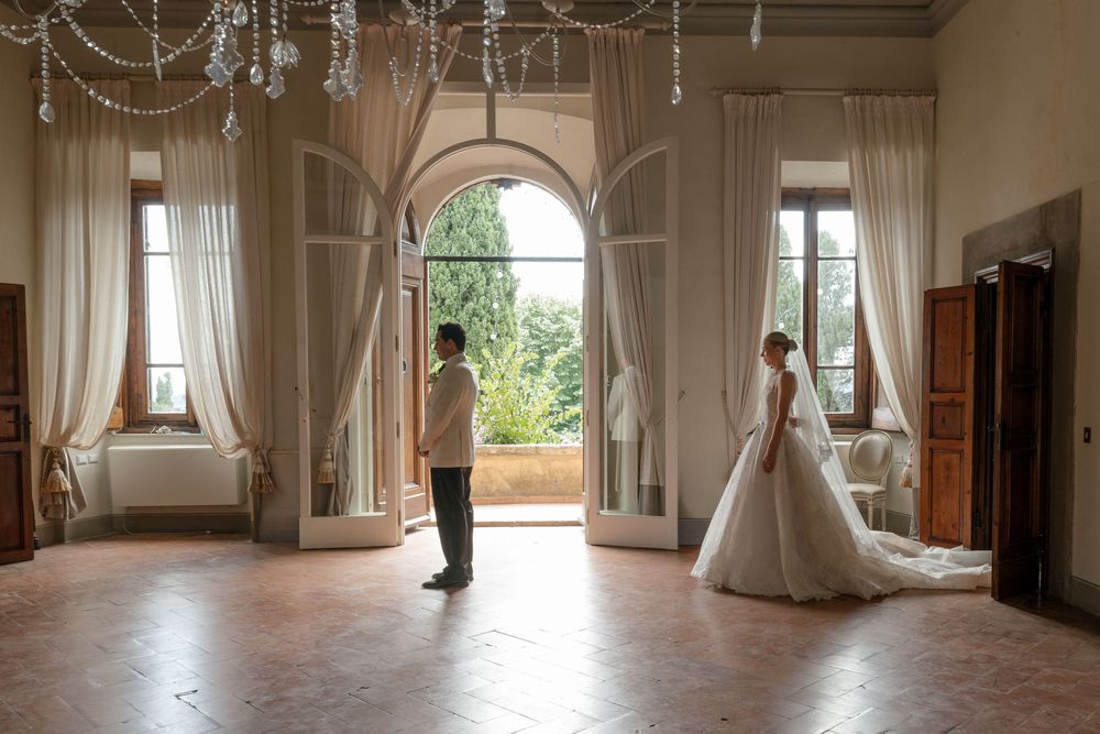 Bride and groom share a romantic first look moment in an elegant villa ballroom with arched doors and flowing white curtains.