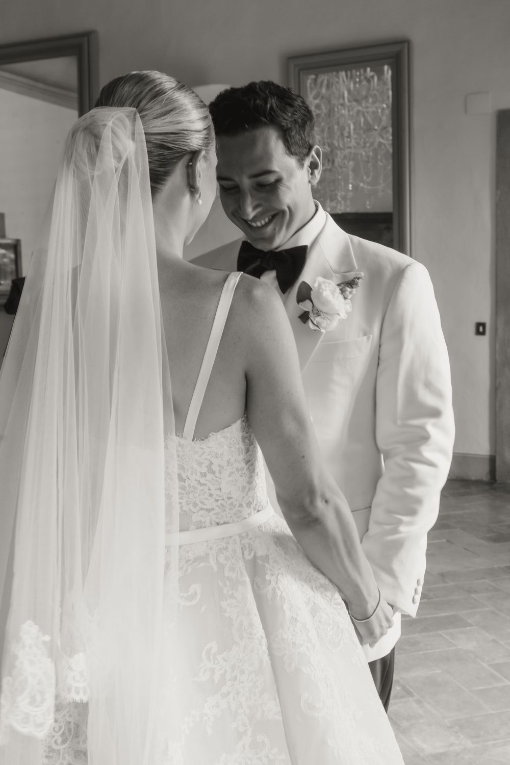 Black and white photo of smiling groom in white tuxedo holding hands with bride in lace wedding dress and veil.