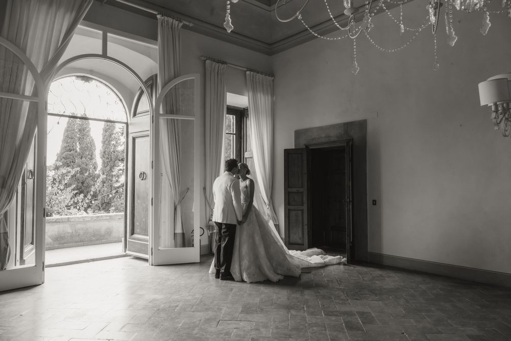 Black and white photo of a bride and groom embracing near elegant arched doors in a grand villa ballroom.