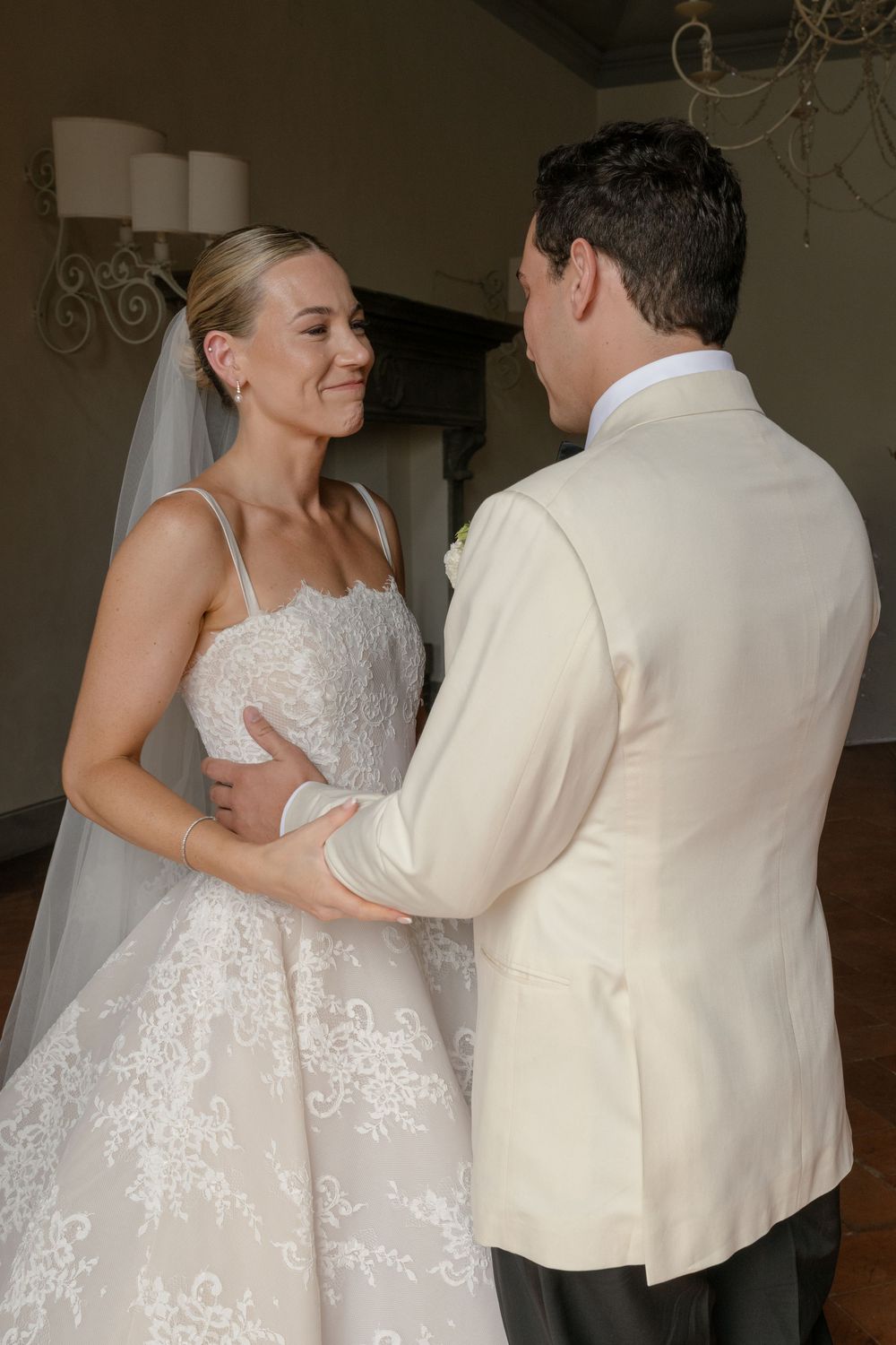 Bride in lace wedding dress and veil smiles at groom in ivory tuxedo jacket during romantic first look.