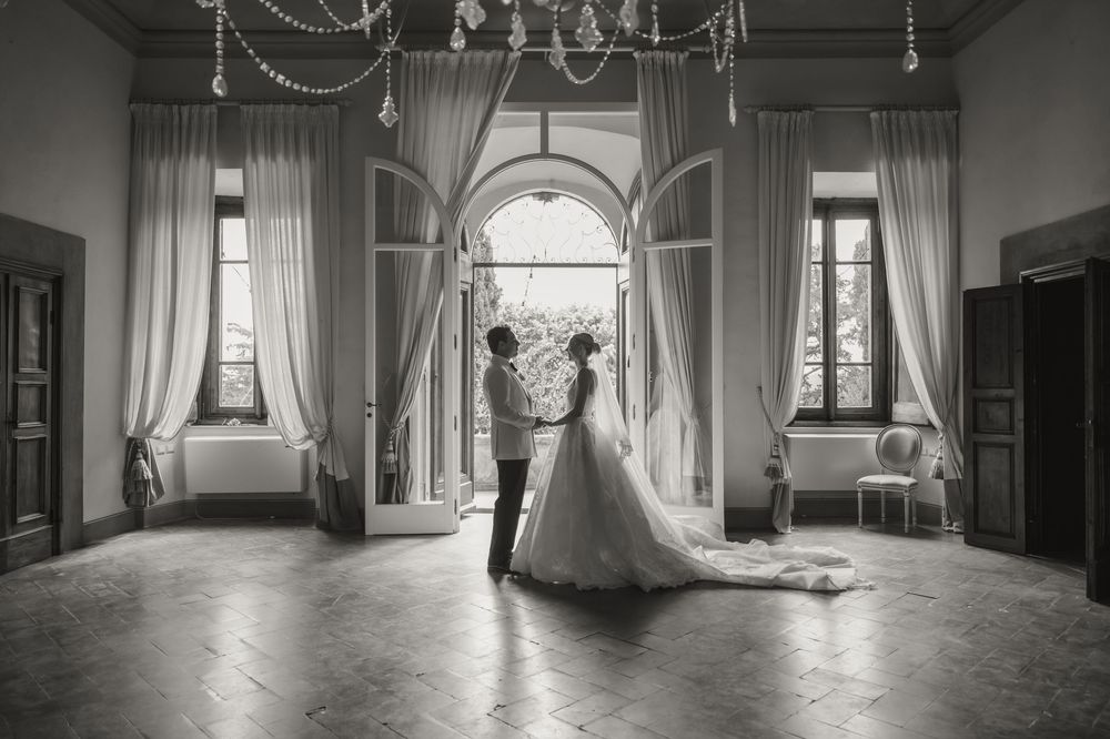 Black and white wedding photo of bride and groom standing in elegant ballroom with grand arched windows and chandelier.