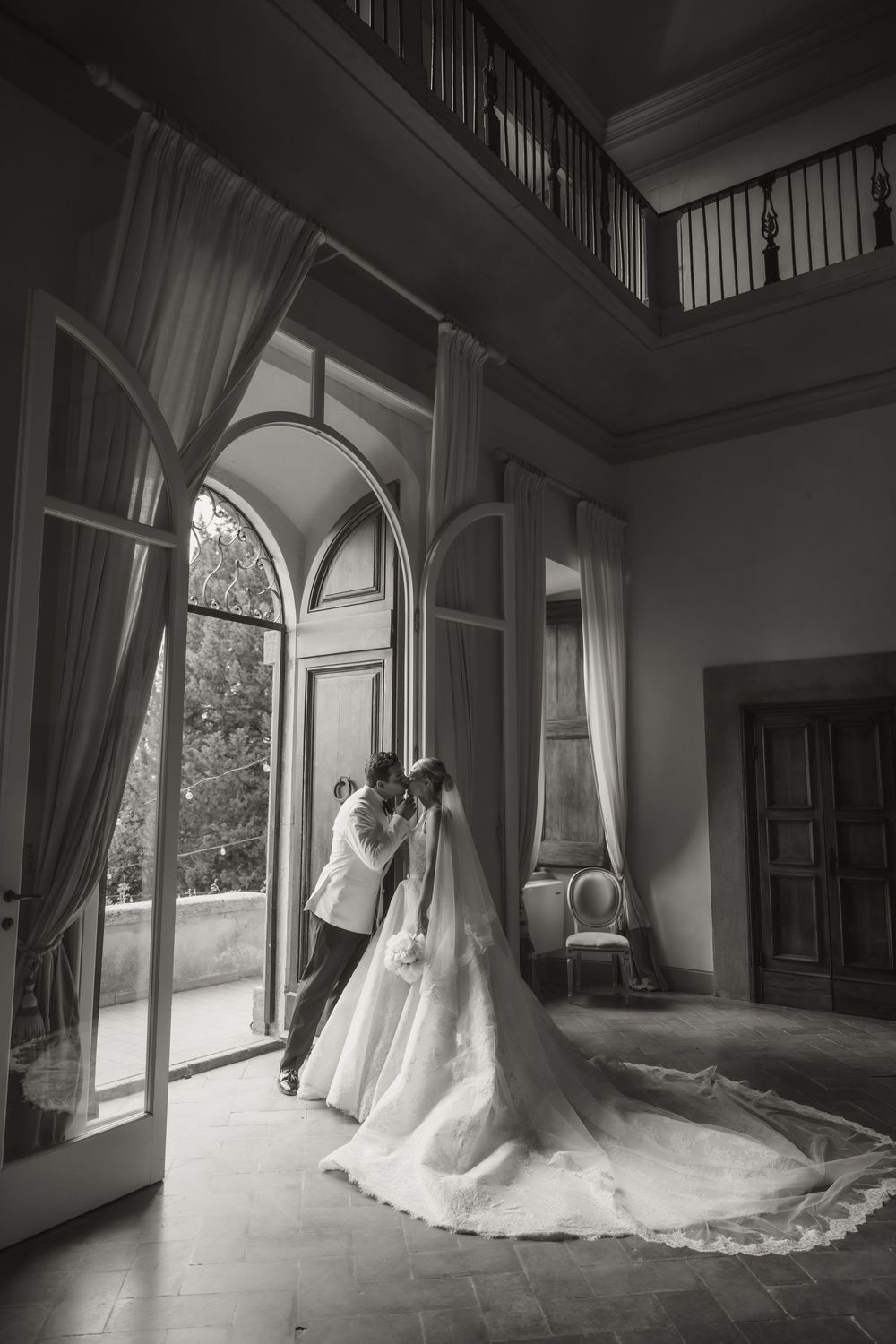 Black and white photo of bride and groom embracing in elegant villa doorway with arched windows and flowing wedding gown.