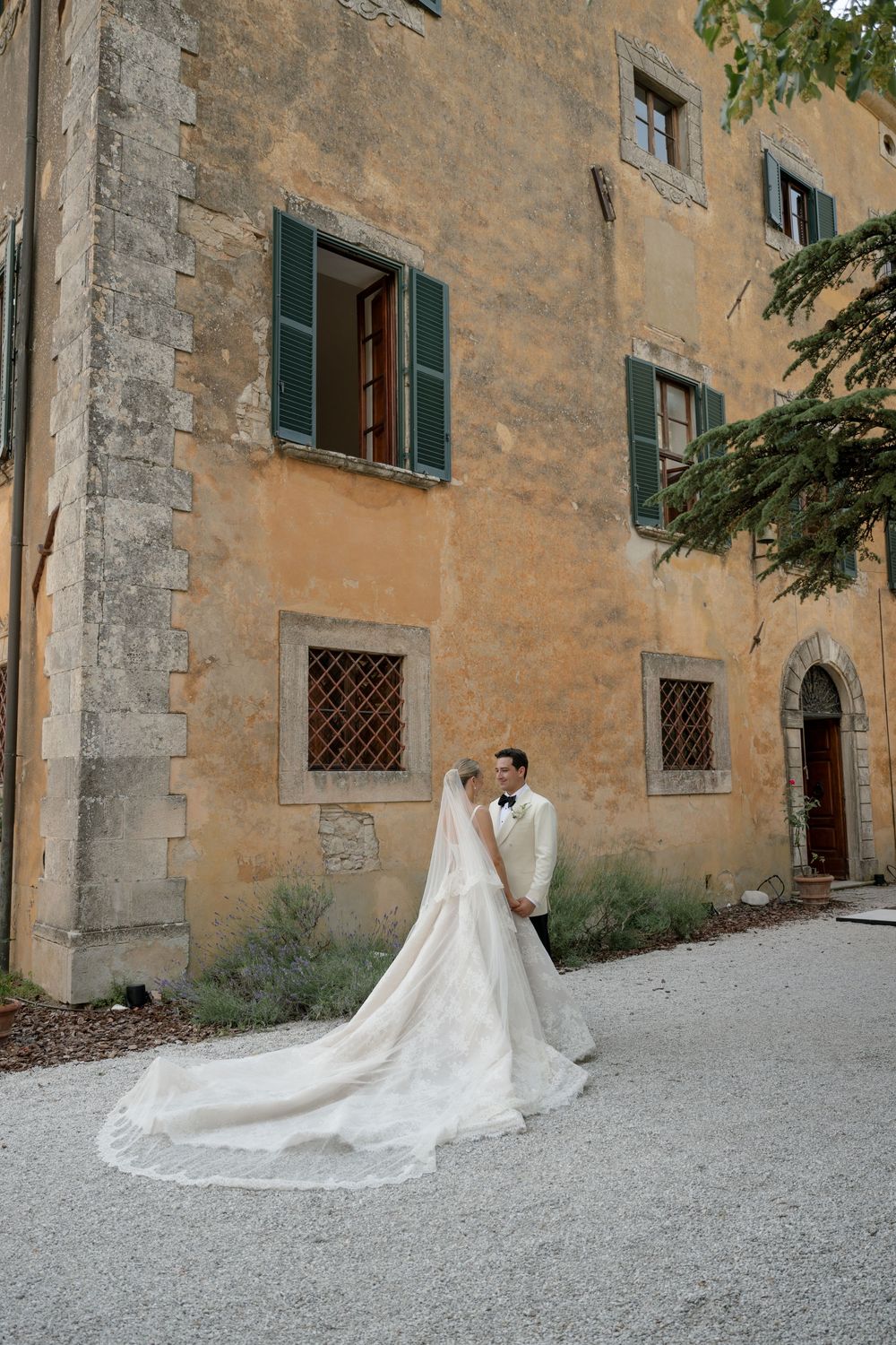 Bride and groom pose before rustic Tuscan villa with green shutters, her long white lace train flowing on cobblestone.