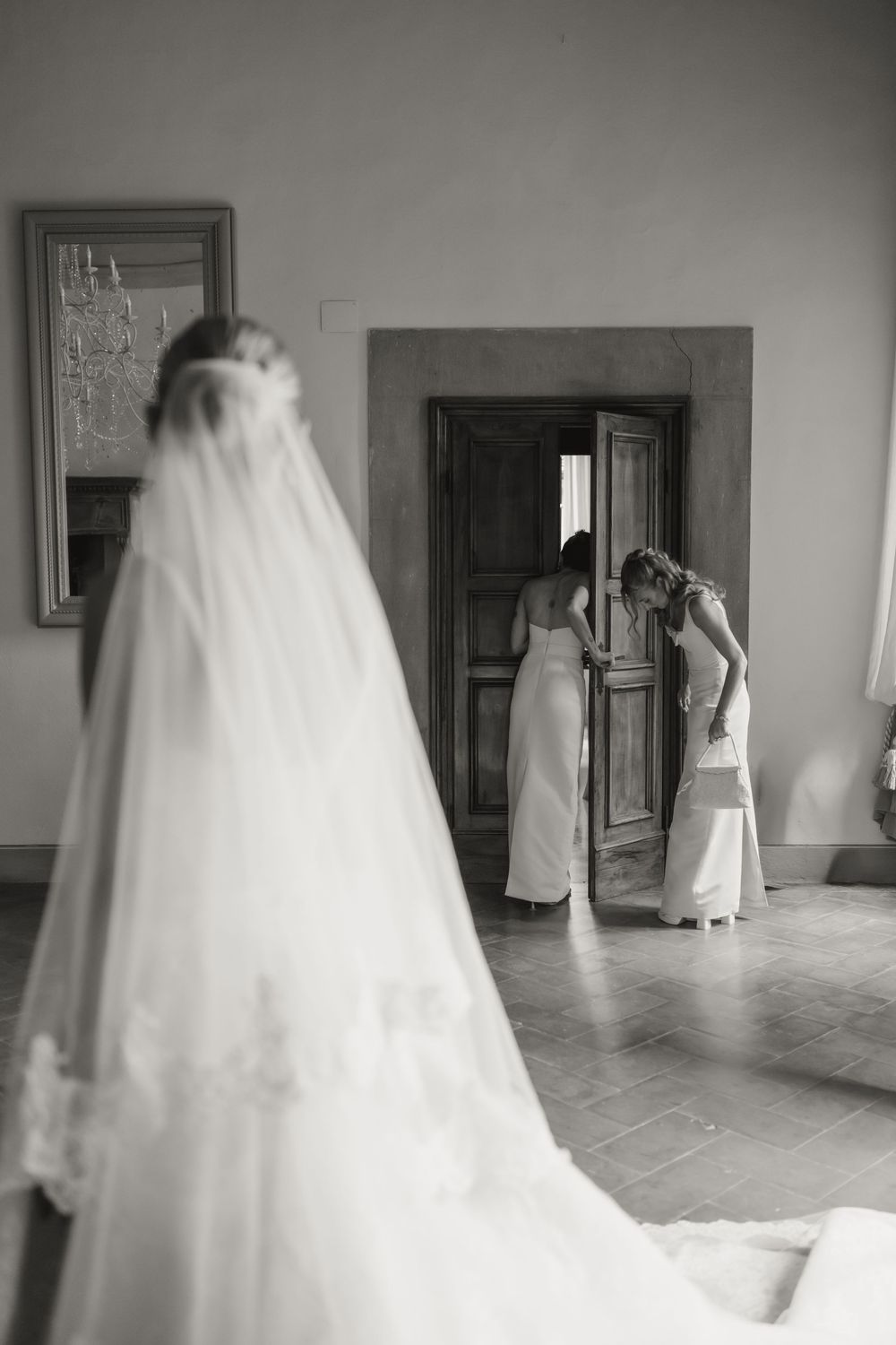 Bride in flowing veil with bridesmaids adjusting wedding dress near ornate doors, elegant black and white photo.