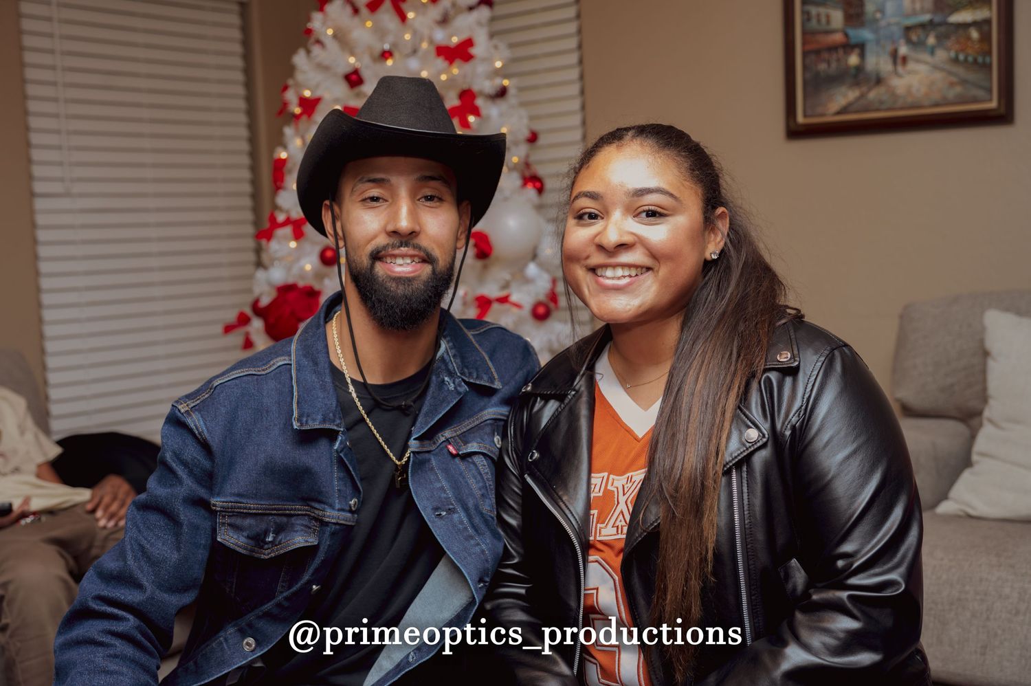 A man in a cowboy hat and denim jacket poses with a woman in a leather jacket near a decorated Christmas tree.