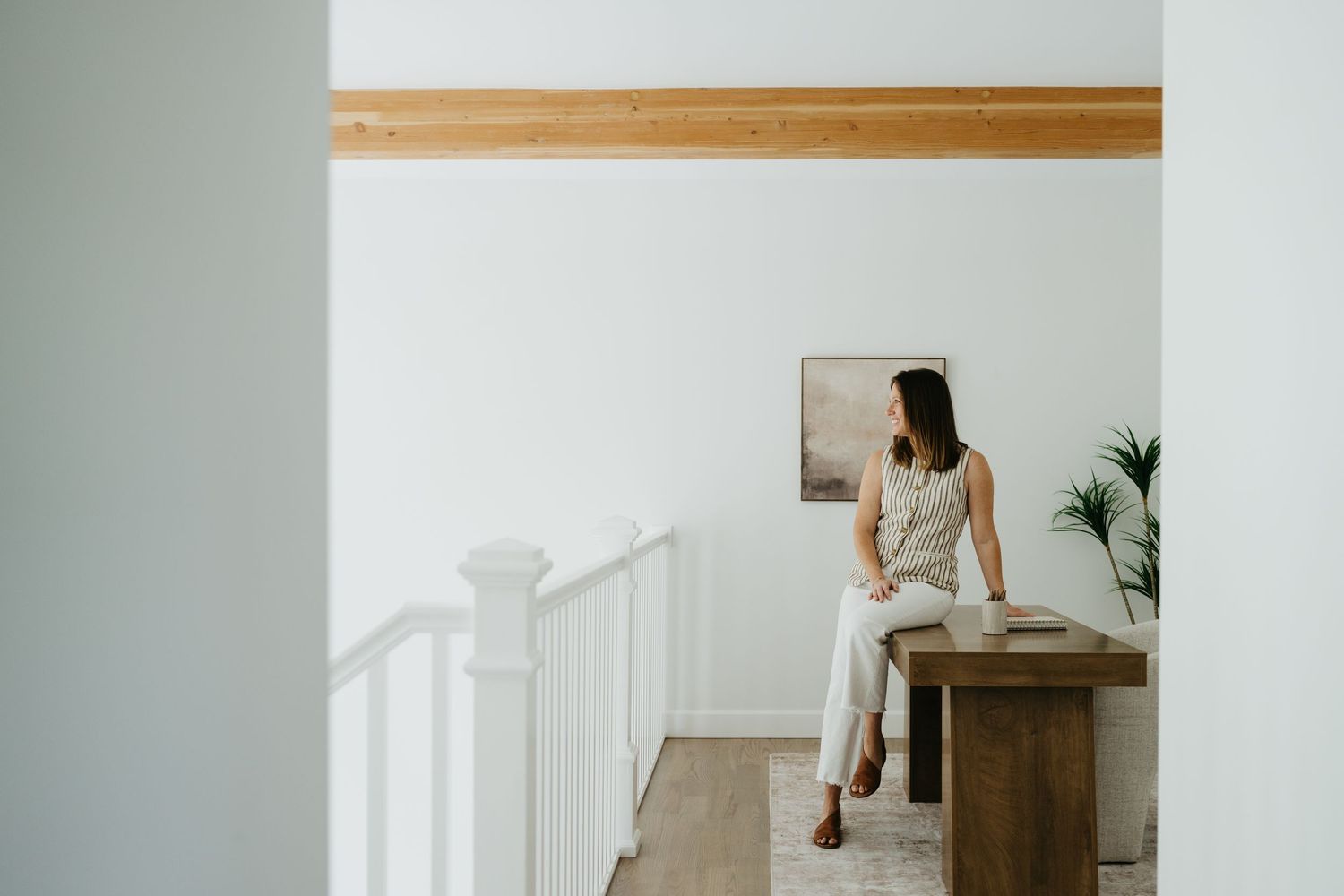 A woman in neutral linen attire sits on a wooden table in a bright minimalist interior near white railings.