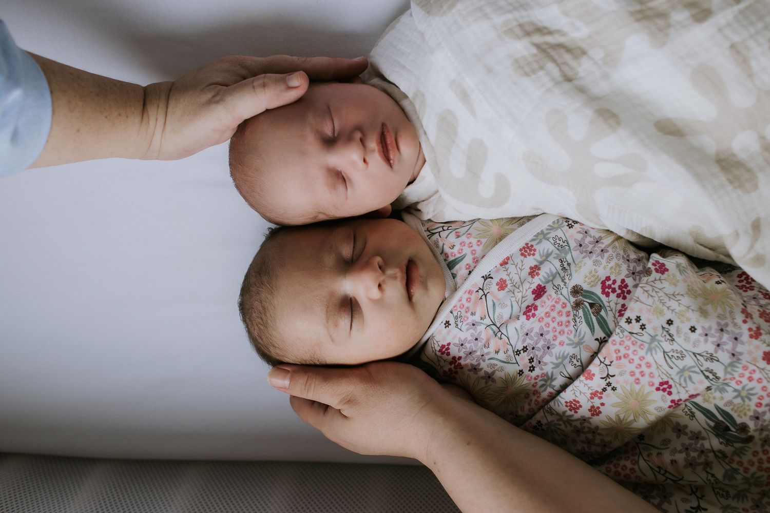 Two newborn twins swaddled and sleeping side by side, gently cradled by a parents hands.