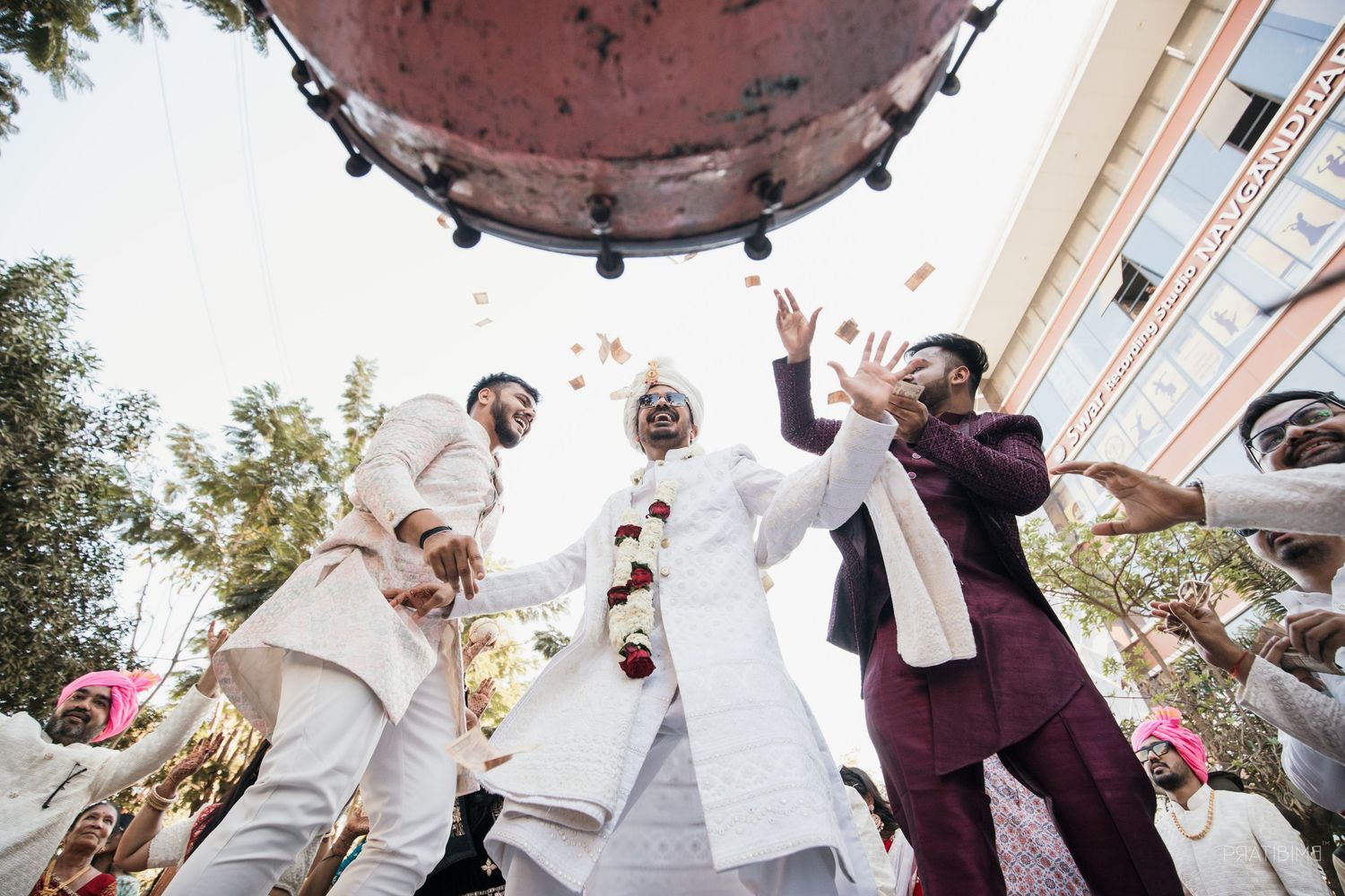 Indian groom in white sherwani celebrates with friends under dhol drum, tossing flower petals at outdoor wedding ceremony.