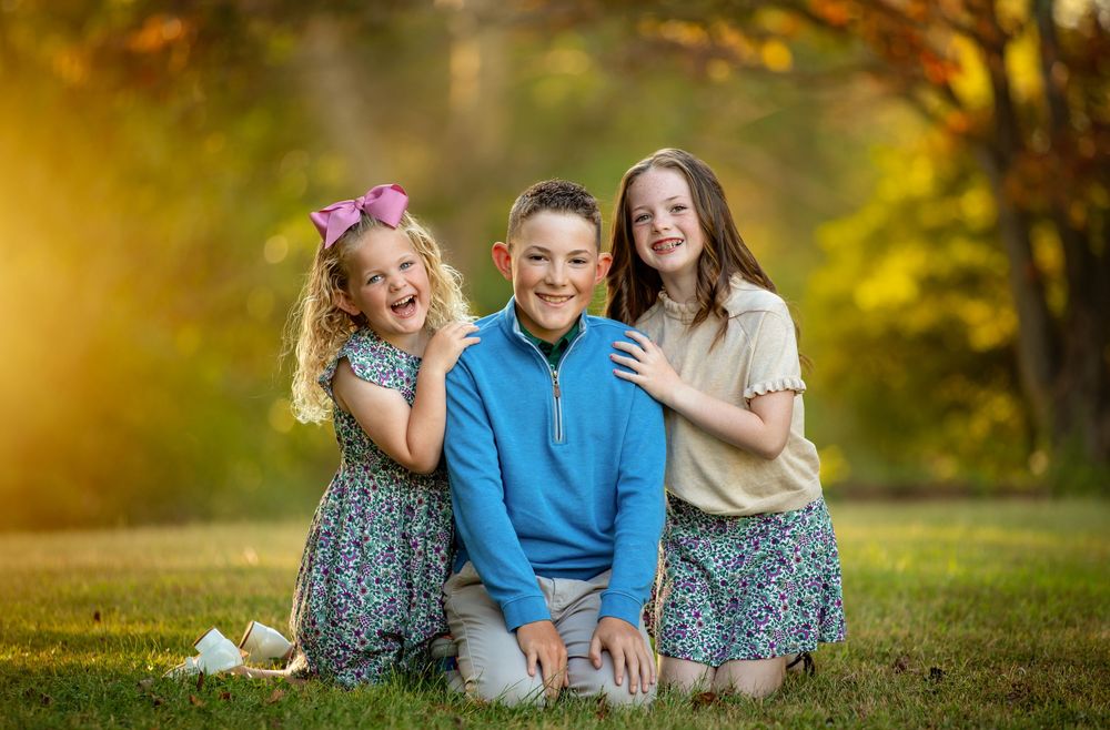 Three children pose together on grass in a park with golden autumn bokeh lighting in the background.