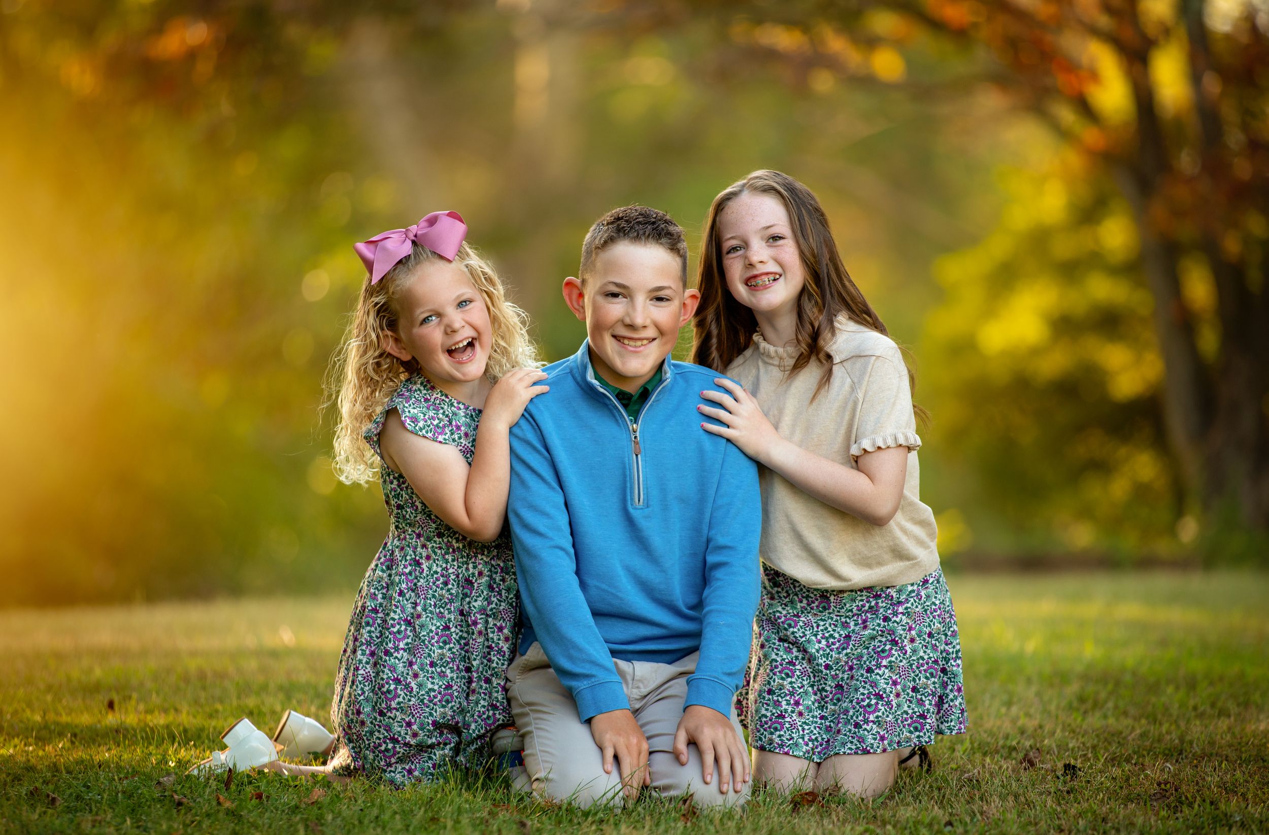 Three children pose together on grass in a park with golden autumn bokeh lighting in the background.