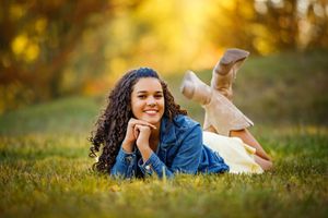Smiling young woman with long curly hair in a denim jacket lies on a grassy field with boots kicked up, glowing in warm golden sunset light.