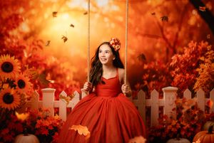 A girl in a red gown sits on a swing surrounded by autumn leaves and sunflowers in a warm fall setting.