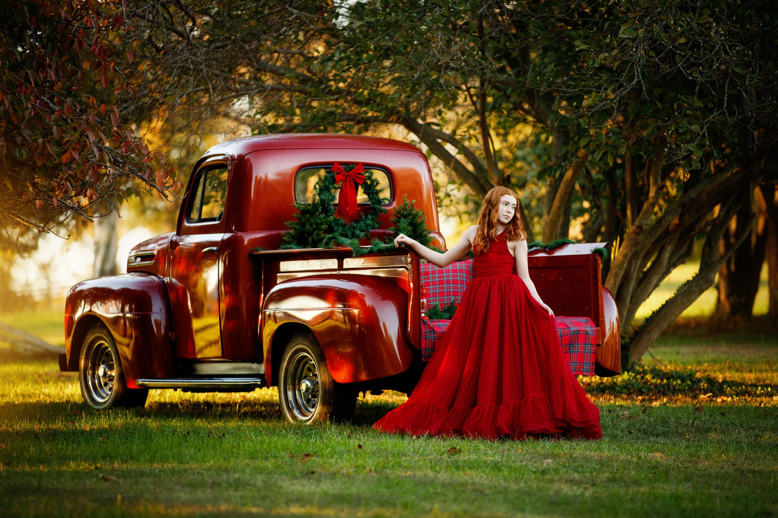 Woman in a flowing red gown stands beside a vintage red pickup truck with a holiday wreath on a grassy lawn under sunlit trees.