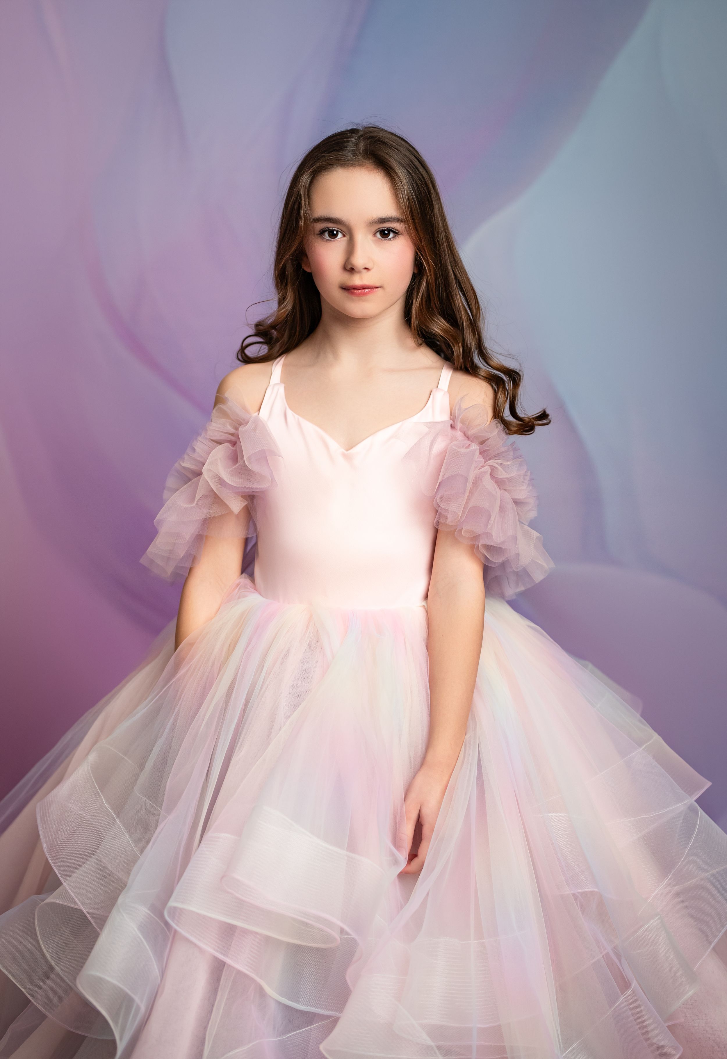 Young ballerina in flowing pink tulle dress poses gracefully against dreamy purple background during dance photoshoot.