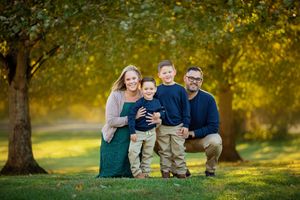 A family group sits together on grass in a sunlit park during autumn with golden light filtering through trees.