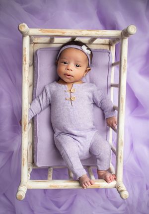 A baby in a purple outfit lies on a purple background while framed by a rustic white wooden frame.
