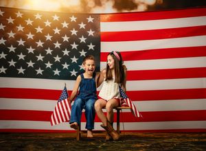 Two joyful girls sitting in front of a large American flag, holding small flags, celebrating Independence Day.