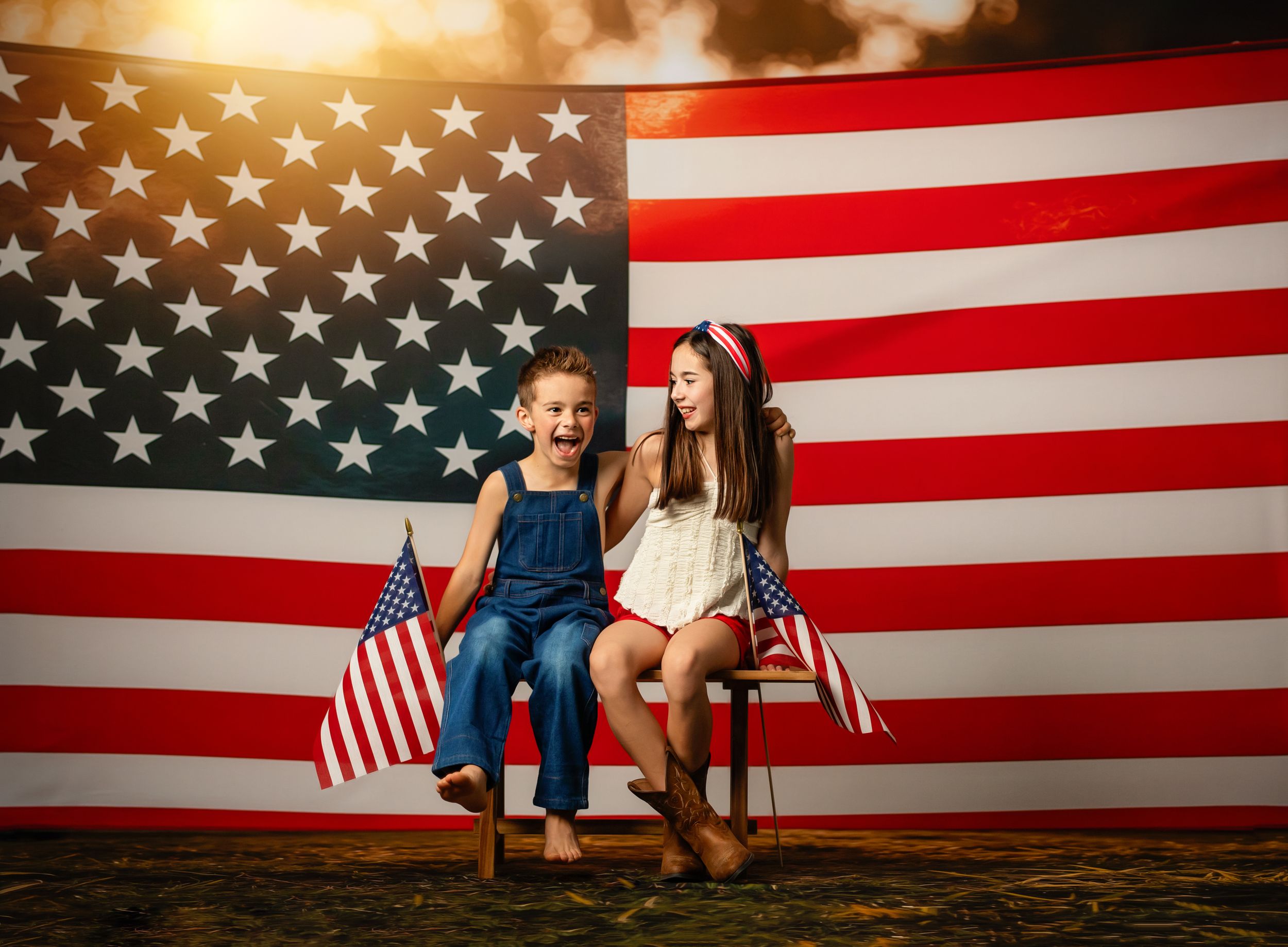 Two joyful girls sitting in front of a large American flag, holding small flags, celebrating Independence Day.