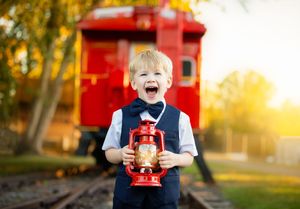 A child in a blue vest laughs joyfully while holding a red toy train during golden hour sunset in a park setting.