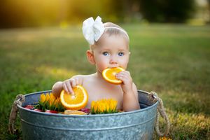 Baby sits in a vintage metal washtub with orange slices and sunflowers creating a sunny summer photo setup.