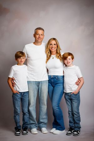 Family of four in matching white tees and jeans posing for a studio portrait photo.
