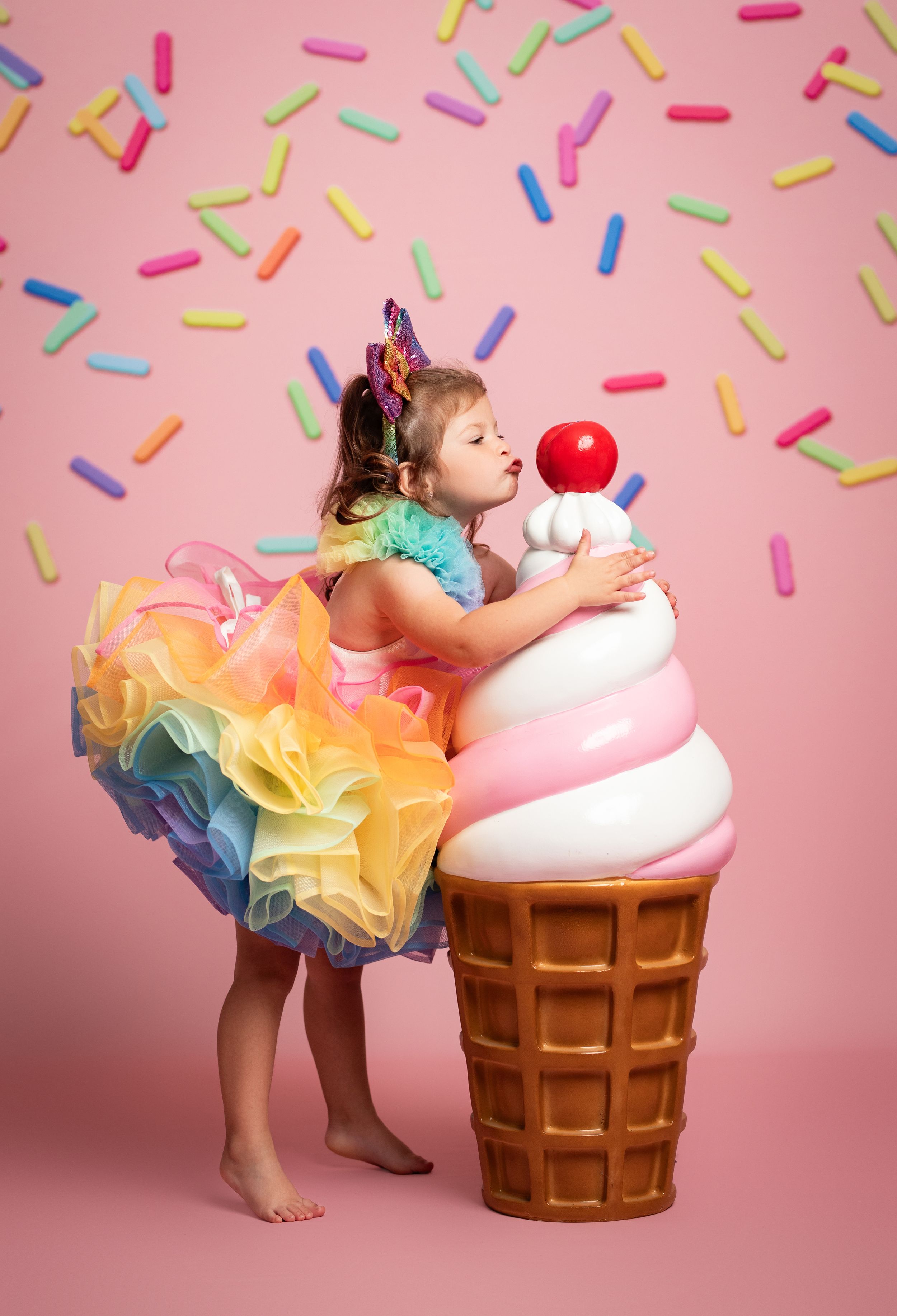 A little girl in a rainbow tutu dress hugs a giant ice cream cone prop on a pink background with colorful sprinkles.