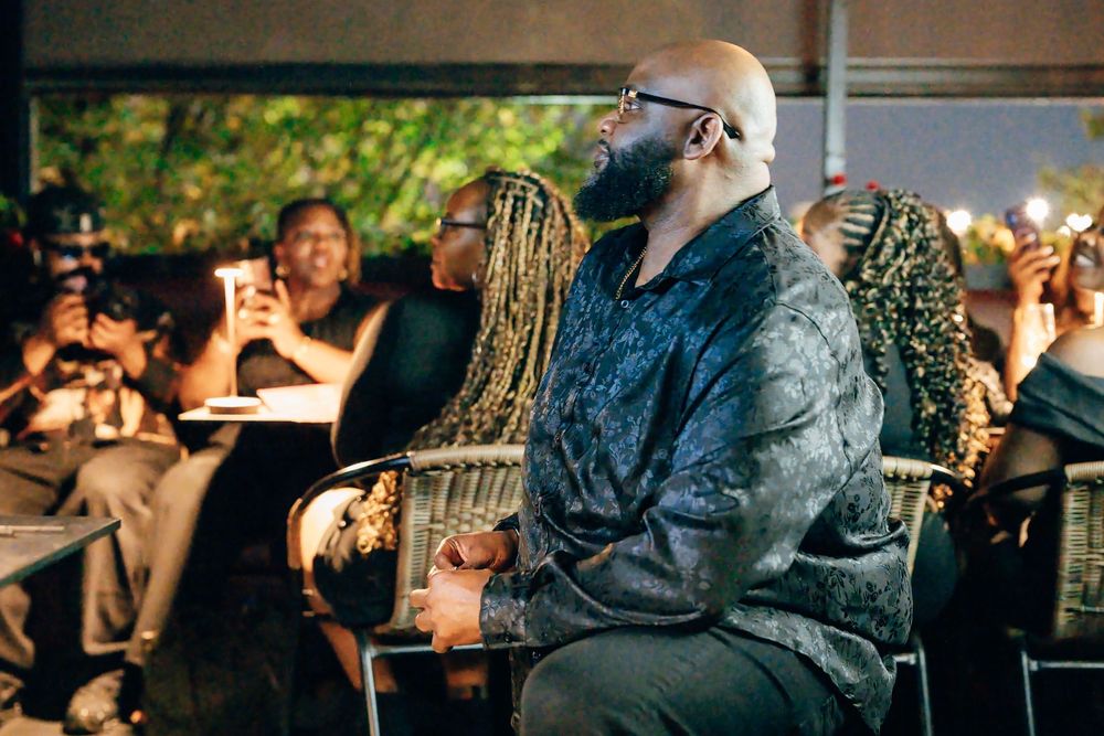 Bald bearded man in glasses and a dark patterned jacket sits in a wicker chair at an evening outdoor event with seated guests behind him.