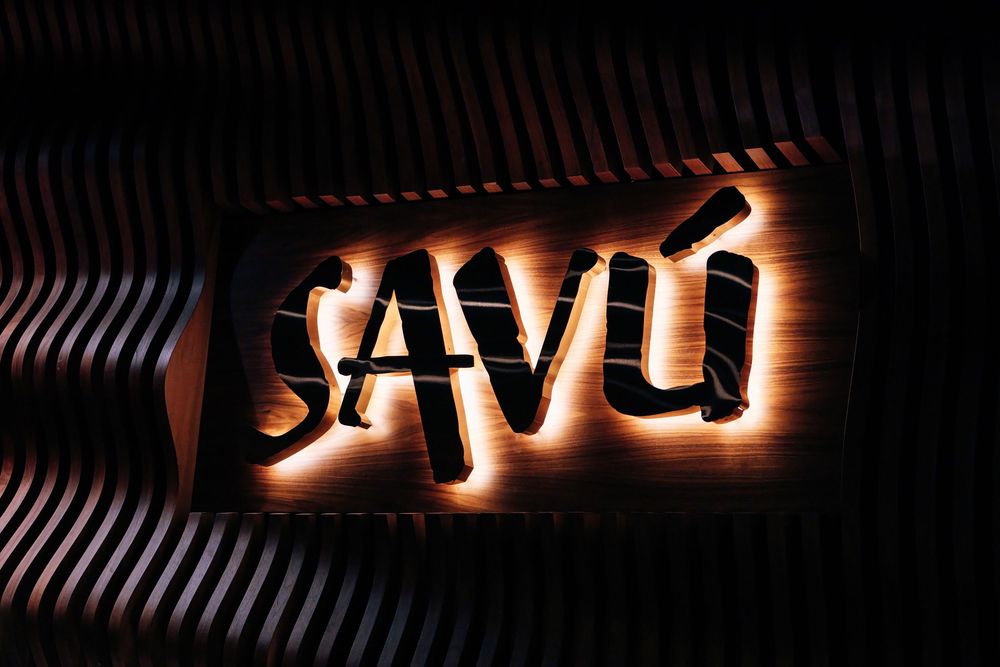 Backlit illuminated SAVÚ sign with warm glowing letters mounted on a dark wall with curved wooden slat paneling.