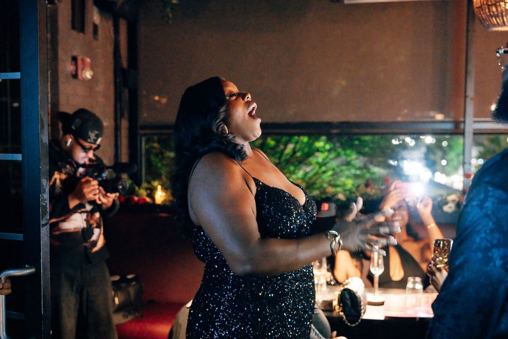 A woman in a sparkling black dress sings passionately with her head tilted back while a photographer captures her at a lively venue.
