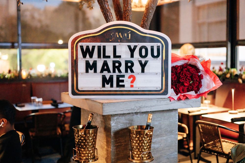 Marquee-style sign reading Will You Marry Me? beside a red rose bouquet and two champagne buckets at a restaurant proposal setup.