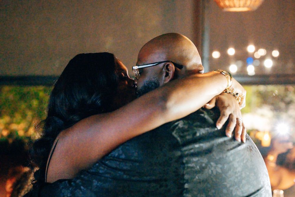 Couple sharing a warm embrace and kiss in a softly lit restaurant, with glowing string lights blurred in the background.