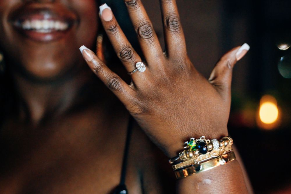 Smiling woman showing off a diamond ring on her raised hand, wearing a stacked gold and gemstone bracelet with a glowing warm background.