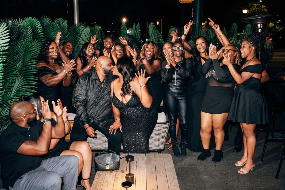 A group of stylish people in black outfits laugh and clap together while gathered around a wooden table at an evening lounge event.